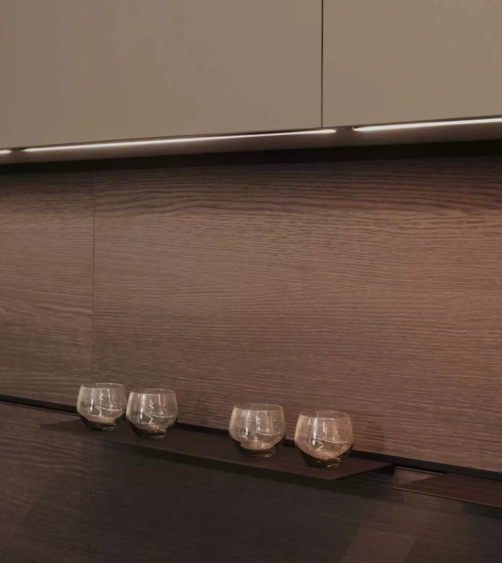 A dark, wood-effect kitchen wall with two-toned cabinetry. The lower section features a rich, dark brown wood grain with a slim black shelf holding four small, rounded glasses. The upper section has taupe/beige cabinetry, and a horizontal LED light strip illuminates the working area below.