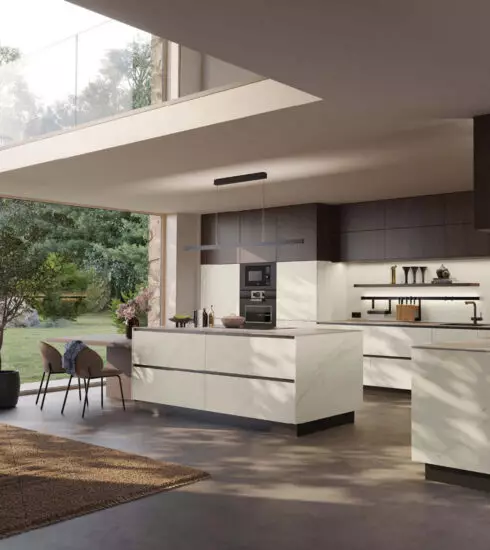 Luxuriöse, moderne Küche mit einer großen Kücheninsel in weißer Marmoroptik. Die Hängeschränke sind dunkelbraun gehalten. Die Küche ist durch eine breite Glasfront zum üppig grünen Garten hin geöffnet.